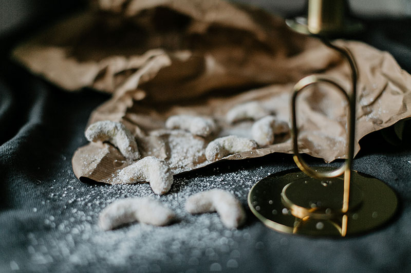 glutenfreie Vanillekipferl liegen zum Teil auf einen grauen Tischdecke, zum anderen Teil auf einem braunen Packpapier. Daneben steht ein goldener Kerzenständer mit einer weißen Kerze.