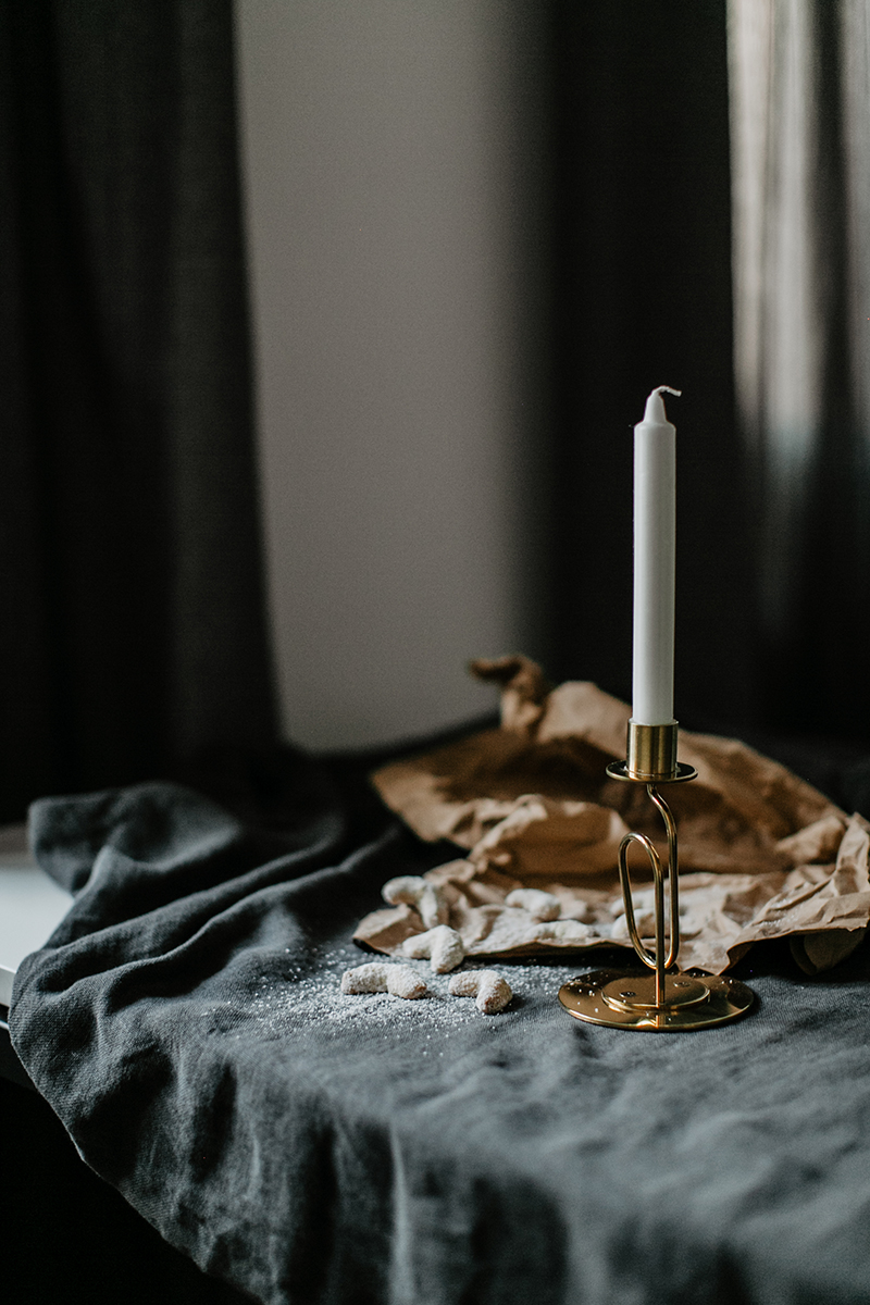 Vanillekipferl liegen zum Teil auf einen grauen Tischdecke, zum anderen Teil auf einem braunen Packpapier. Daneben steht ein goldener Kerzenständer mit einer weißen Kerze.